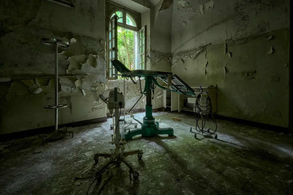Sala operatoria abbandonata con lettino chirurgico, pareti deteriorate e luce naturale proveniente da una finestra aperta.