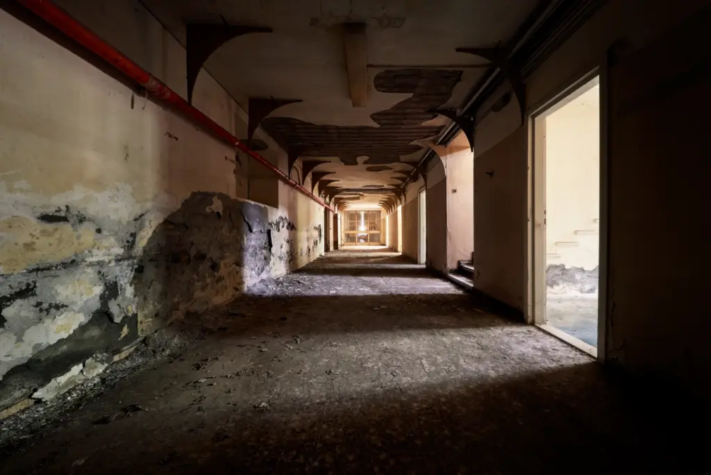 Lungo corridoio di un edificio abbandonato con arcate, luce naturale laterale e prospettiva centrale.