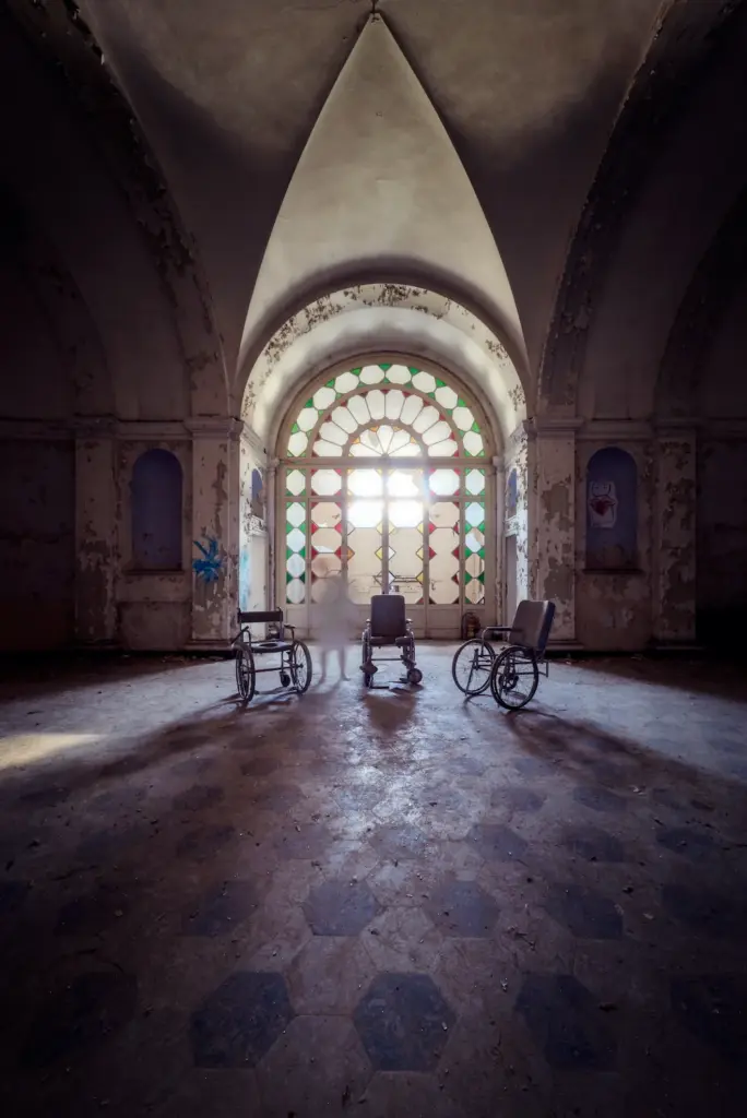 Sala di un manicomio abbandonato con tre sedie a rotelle davanti a una grande vetrata colorata, illuminata dalla luce naturale